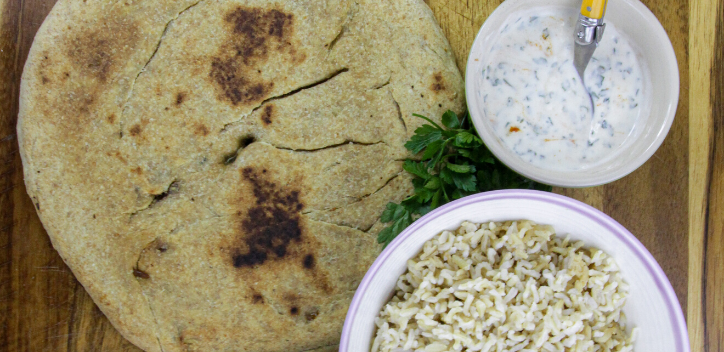 A filled flatbread served on a wooden board, with a small bowl of soya yoghurt dressing, and a bowl of rice served on the right hand side.