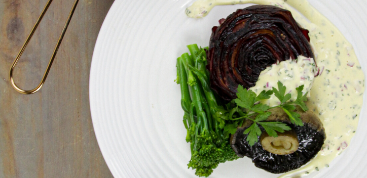 A beetroot stake served with stem broccoli, flat mushrooms and a soya cream sauce on a round plate.