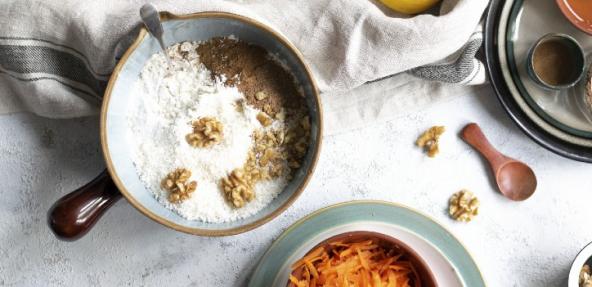 Flour, cinnamon and walnuts in a mixing bowl next to a bowl of grated carrots.