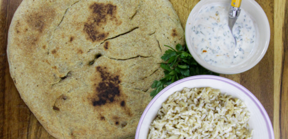 A filled flatbread served on a wooden board, with a small bowl of soya yoghurt dressing, and a bowl of rice served on the right hand side.