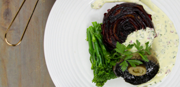 A beetroot stake served with stem broccoli, flat mushrooms and a soya cream sauce on a round plate.
