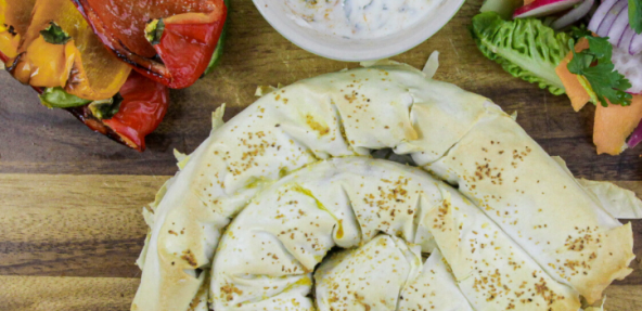 A filled pastry swirl served on a wooden board with mixed peppers, veg and soya yoghurt dipping to the side.