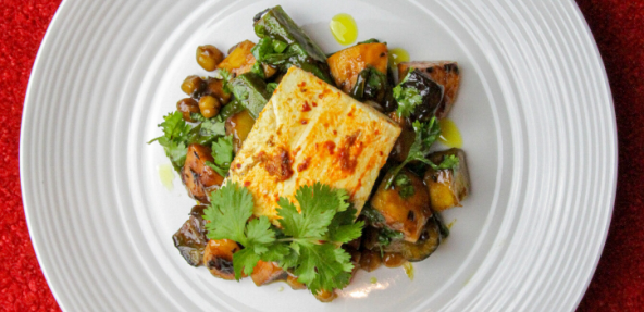 Vegan Brinjal pickle vegetables and spiced tofu steak with a green garnish on a white plate against a red background