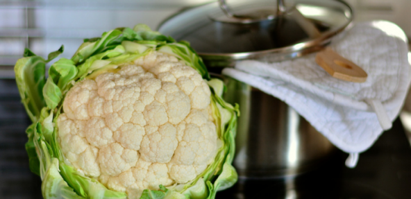 A cauliflower head resting against a steel saucepan with a tea-towel and wooden spoon draped over the top.