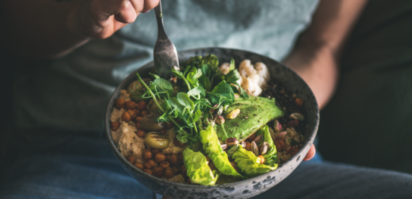 Healthy dinner or lunch. Person in t-shirt and jeans eating vegan superbowl or Buddha bowl with hummus, vegetable, salad, beans, couscous and avocado