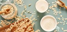 Oat milk in a glass and mug on a blue background. Flakes and ears for oatmeal and granola on a wooden plate.
