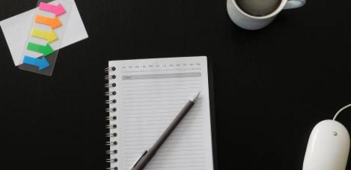 A writing pad with a pen resting on top.  A mug of coffee to the top right, a computer mouse bottom right and a row of note arrows top left all against a dark background.