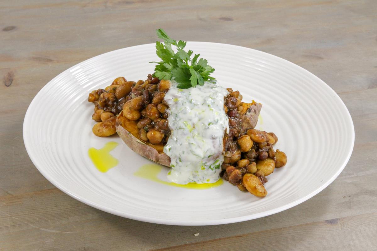 vegan lentil and bean chilli on top of a baked sweet potato with creamy sauce over the top and a green garnish