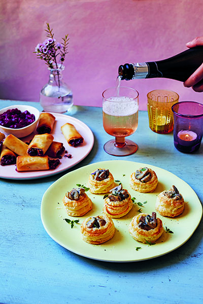 Mushroom canapes on a plate on a blue table with another plate of food in the background to the left, to the right is a drink and two candles