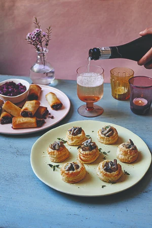Mushroom canapes on a plate on a blue table with another plate of food in the background to the left, to the right is a drink and two candles