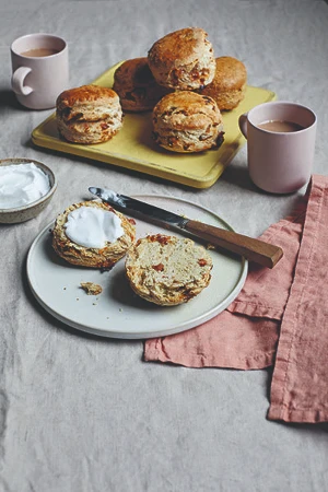 Batch of scones on a tray. One scone sliced up on a plate with a knife next to it, on top of a tea towel with two cups of tea