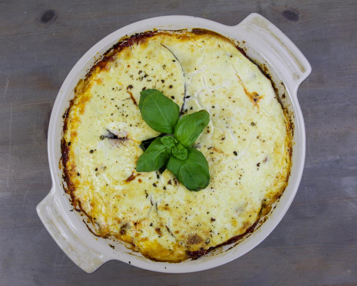 vegan lentil and tofu moussaka in a white round oven dish with basil garnish on a grey background