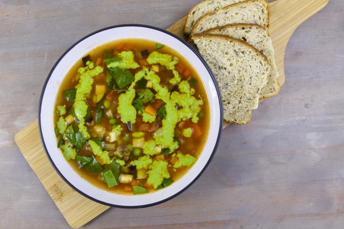 vegan seasonal minestrone with green lentils and pistou in a bowl with a blue edge on a wooden board with slices of brown bread against a grey background