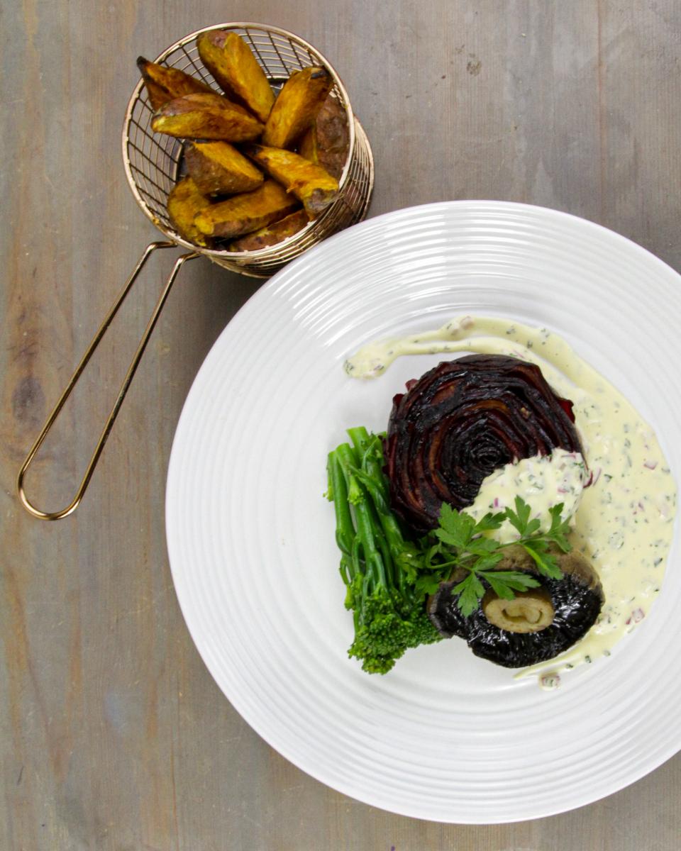 vegan beetroot steak with Bearnaise-style sauce on a white plate with tenderstem broccoli and mushrooms on a grey surface with a small metal basket of wedges 