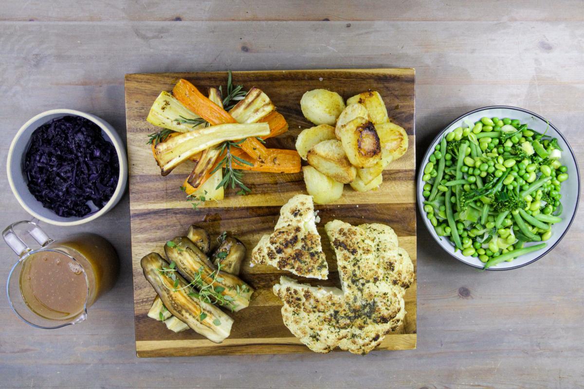 vegan roast cauliflower Sunday lunch deconstructed on a wooden board with a jug of gravy and a separate bowl of peas and broccoli