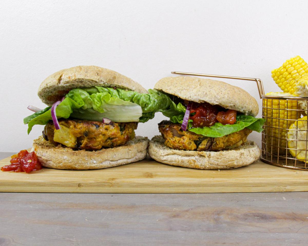 two leftovers burgers on a wooden board with a small basket of corn on the cob against a grey background