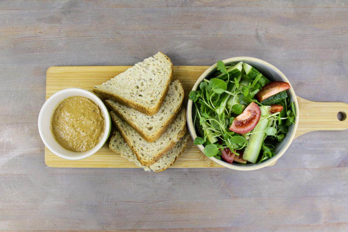 vegan lentil and mushroom pâté in a small white bowl with slices of brown bread next to it and a small bowl of salad next to the bread all on a wooden board against a grey surface