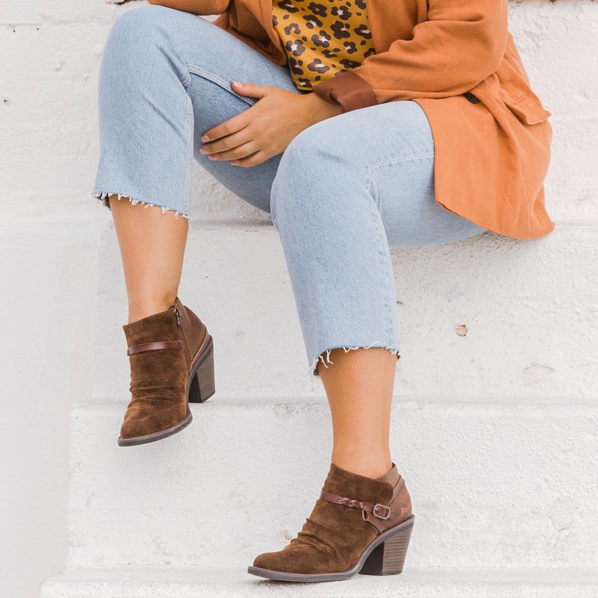 Female sitting down image from chest down showing brown top, blue jeans and brown ankle boots