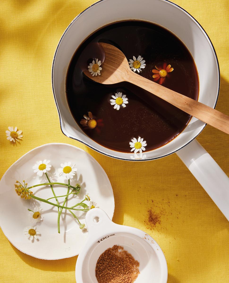 A milk pan filled with a dark brown mixture of coconut sugar syrup, decorated with chamomile flowers, resting against a bright yellow table top.