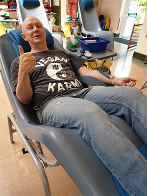 David Harriman doing a thumbs up to the camera, laying down, in the process of giving blood.