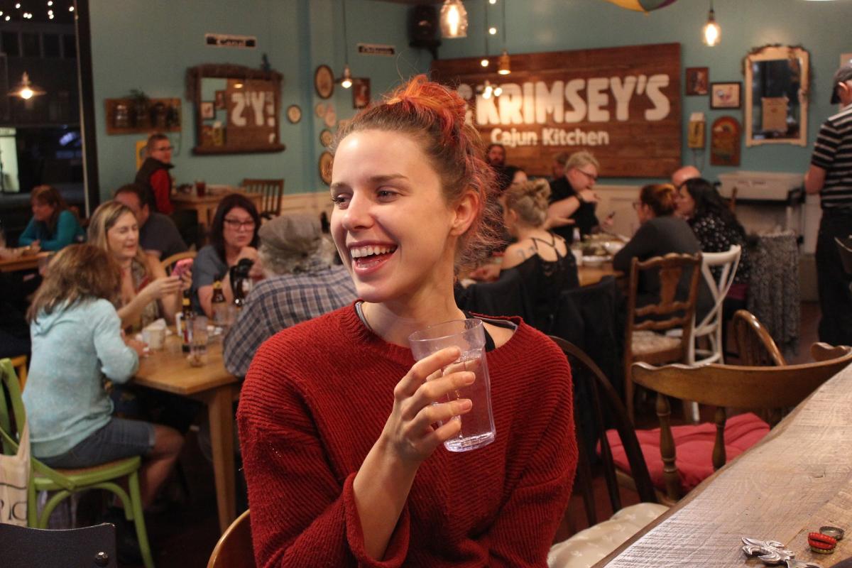 Girl sitting and smiling at Krimsey's Cajun Kitchen bar with drink in her hand and view of the restaurant behind her