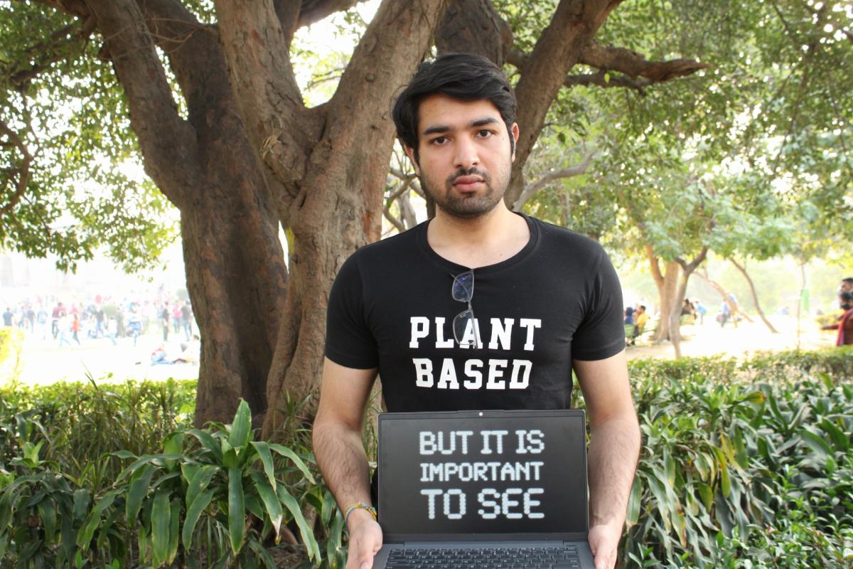 Image if Ishaan outside wearing a plant based t-shirt, holding a laptop reading 'but it is important to see' written on it.