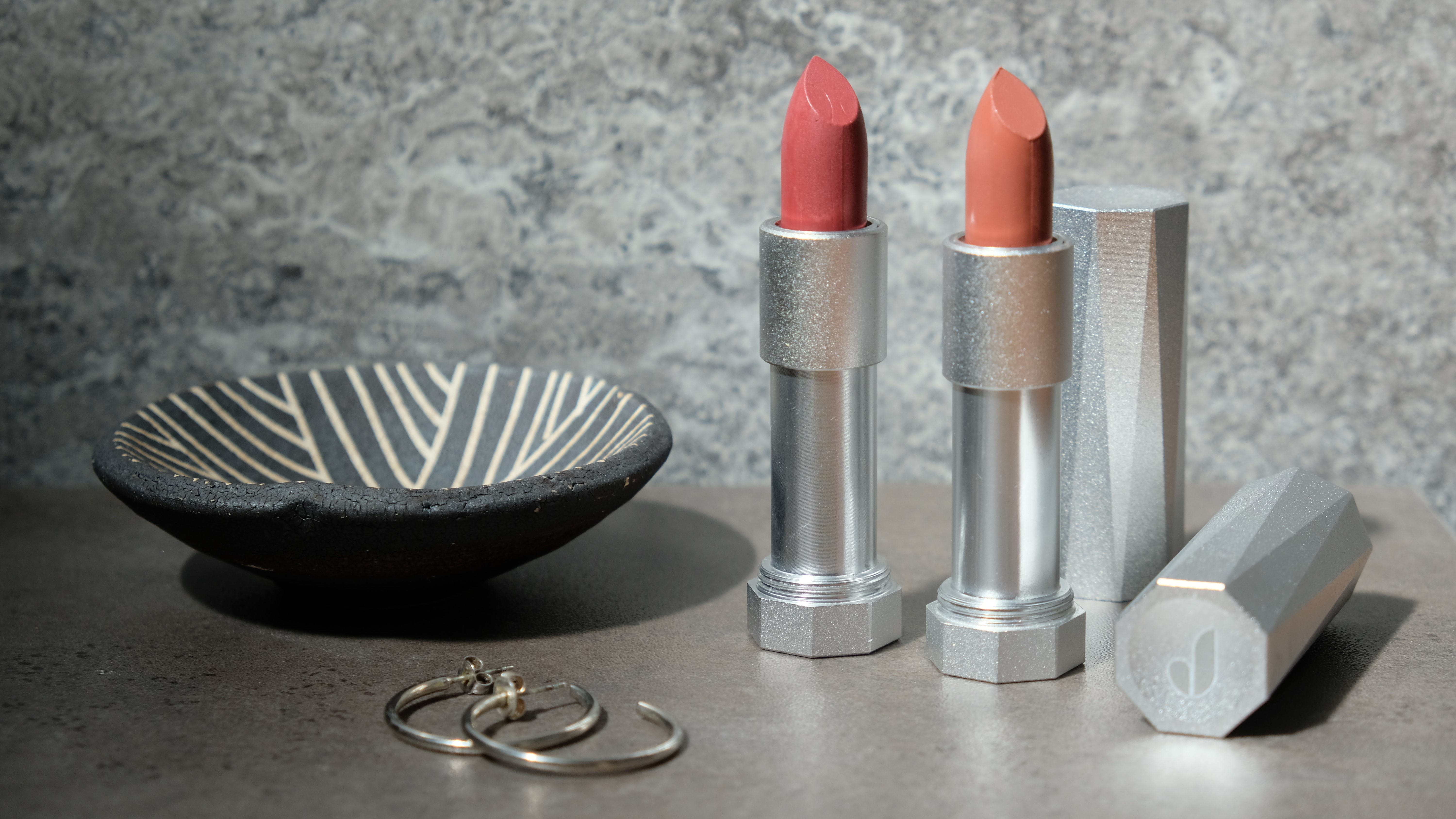 Two Juni lipsticks in pink and nude against a marbled background with a bowl and earrings 