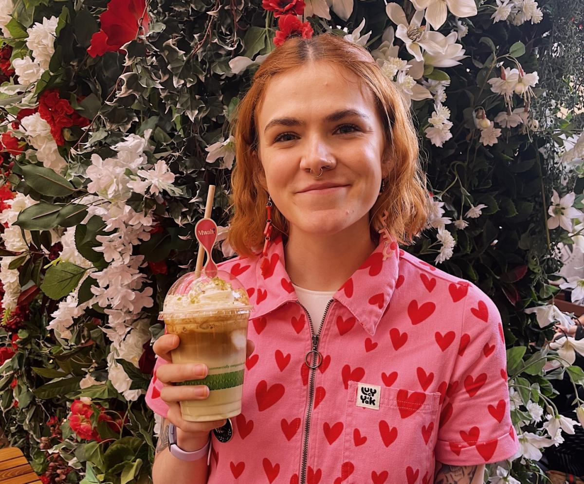 Vegan LGBTQIA+ influencer Kelsey, standing against a flower backdrop with a vegan drink
