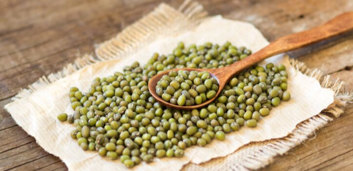 Mung beans in a wooden spoon on a piece of woven cream cloth with frayed edges on a wooden table