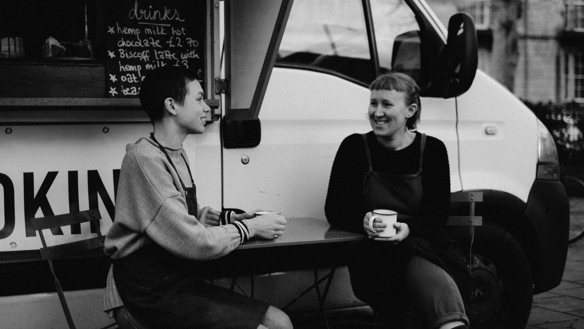 Black and white image of two women sat outside of a vegan food van image talking and smiling