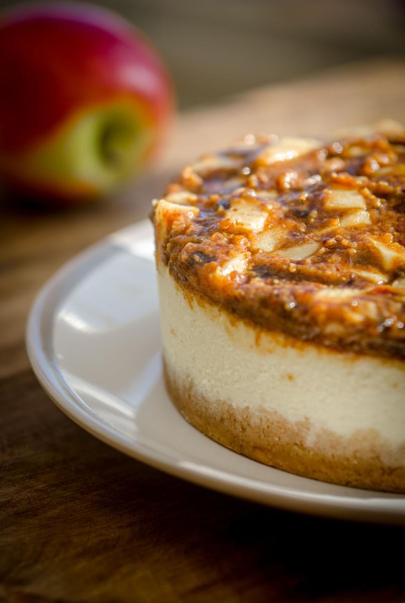 A zoom in of a cheescake on a plate, topped with apples and caramel.