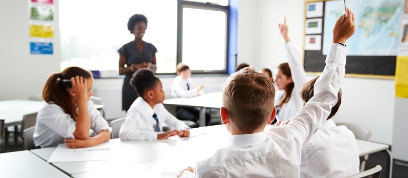 High School Students Wearing Uniform Raising Hands To Answer Question Set By Teacher In Classroom