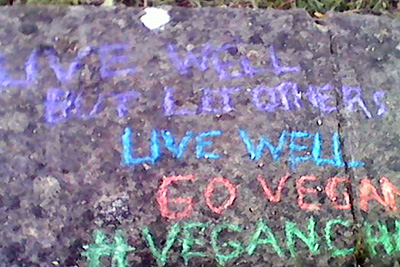Chalk message on pavement reading "live well but let others live well. Go Vegan #veganch" the message is cut off.