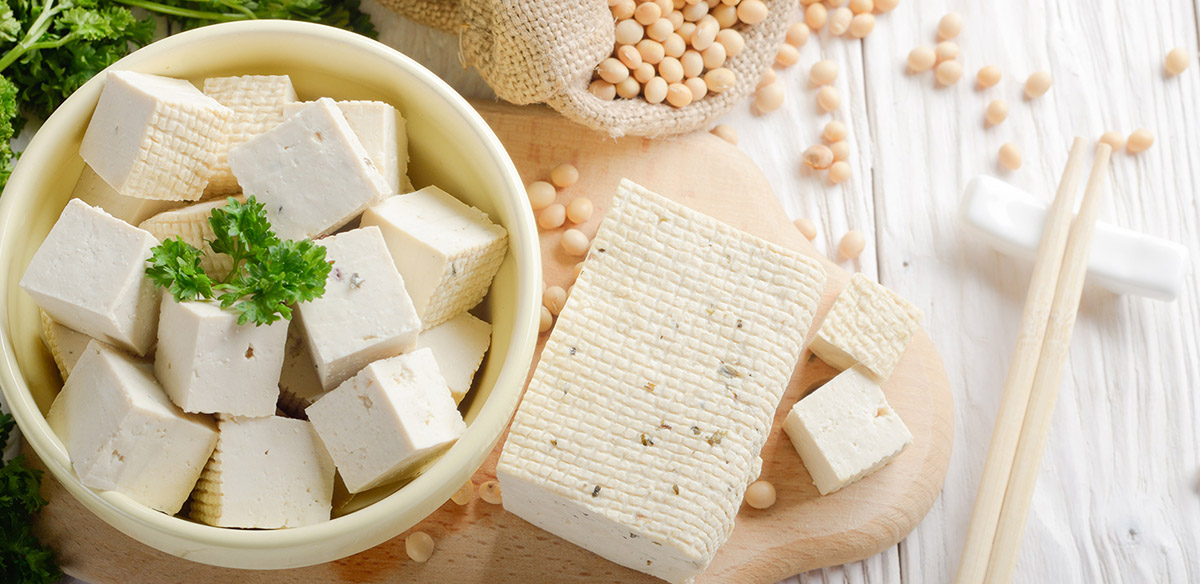 A photograph of a block of firm tofu on a chopping board, along with a small pot with cubes of already chopped up tofu. Around this are scattered soya beans and greens.