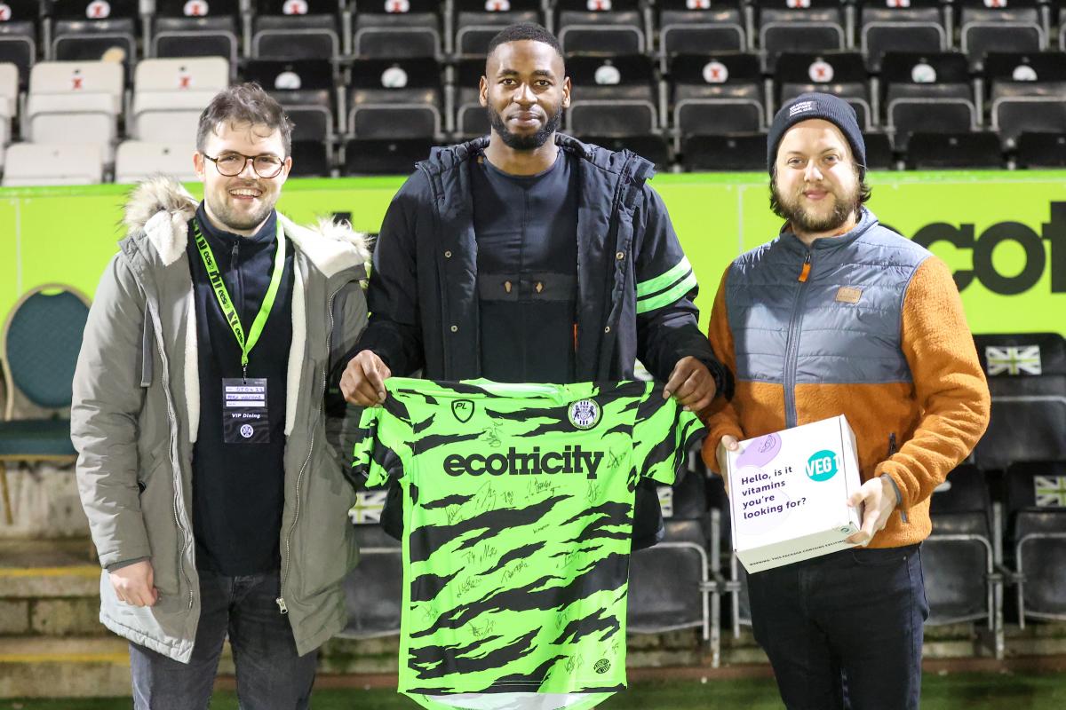 Three men from Forest Green Rovers football team, standing in a stadium holding an Ecotricity shirt